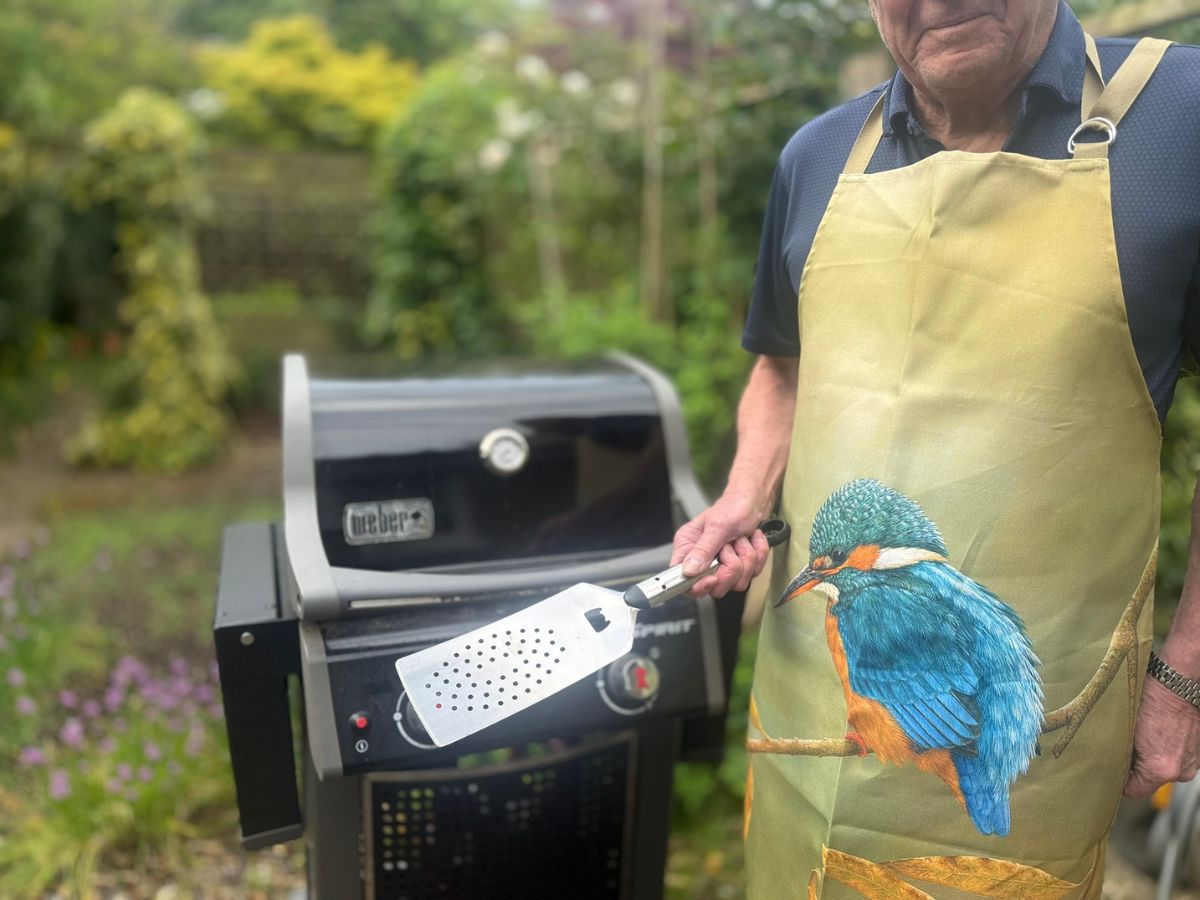 man wearing kingfisher apron in front of bbq holding spatula