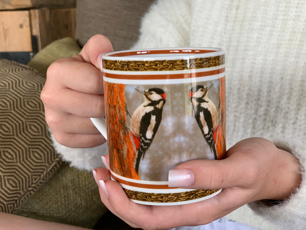 A person holding a fine bone china mug with a woodpecker design against the trunk of a tree, decorated along its inner rim.