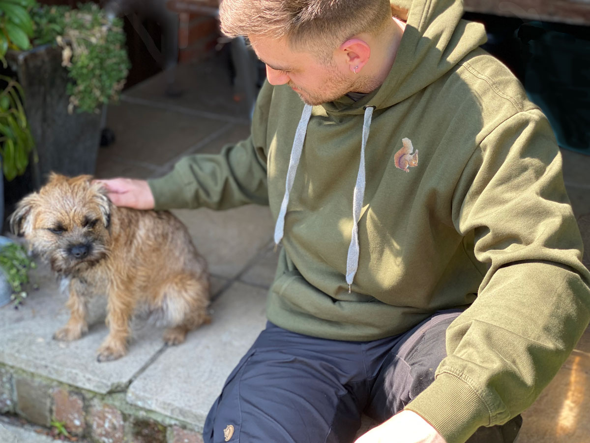 man wearing khaki coloured hoodie with squirrel emblazoned on it petting dog