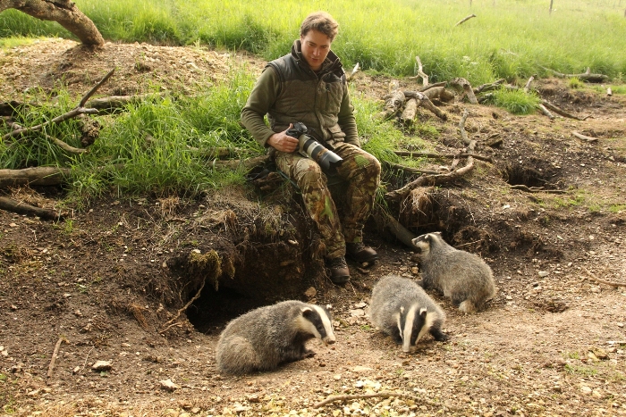 artist Robert E Fuller with camera in hand sitting on edge of badger sett with three cubs at his feet