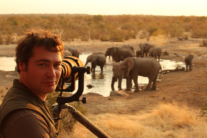 artist robert fuller turned away from camera which is pointing at waterhole with elephants drinking